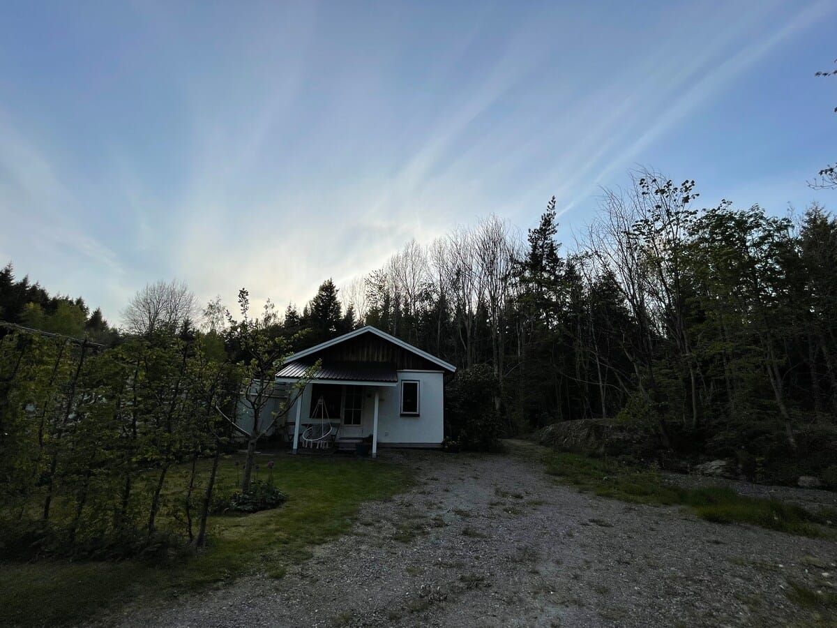 Vita huset i kvällsljus — en låg vit stuga med röd-svart tak, gräsmatta och skog runt om.